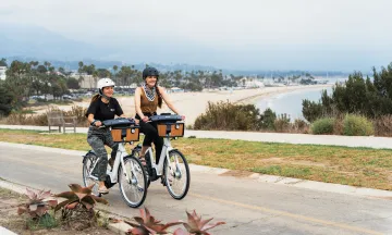 Two women wearing helmets ride BCycle bikes along coast SB
