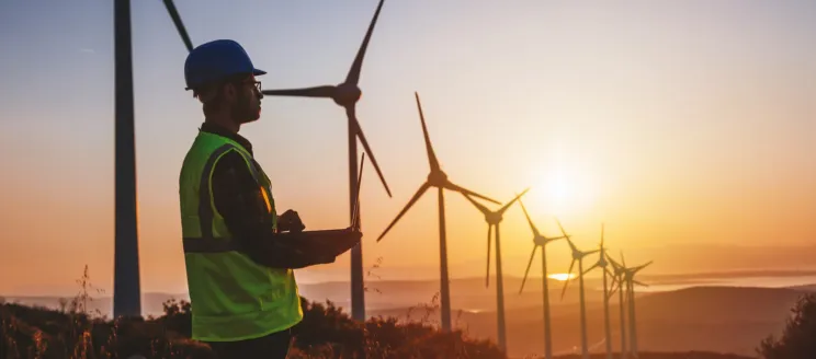 Stock image of a man in safety vest and hard hat in front of wind turbines with sunrise in the distance
