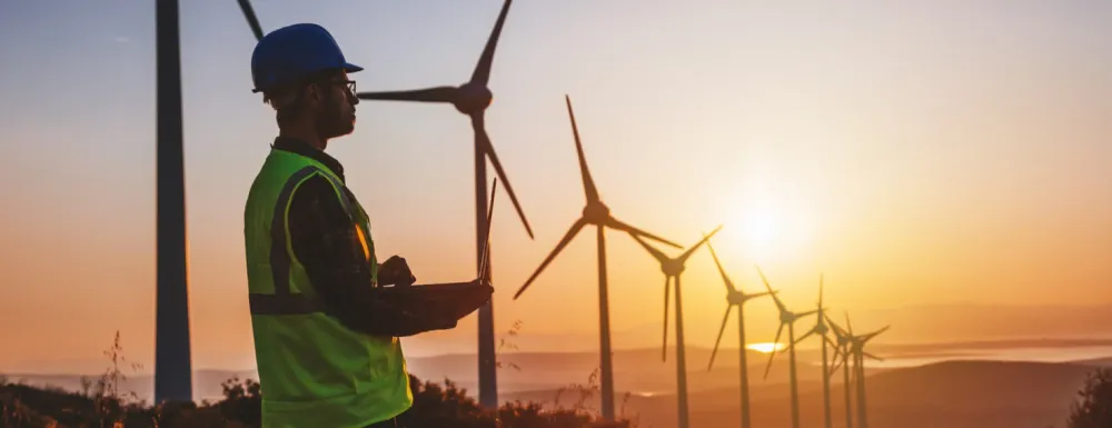 Stock image of a man in safety vest and hard hat in front of wind turbines with sunrise in the distance