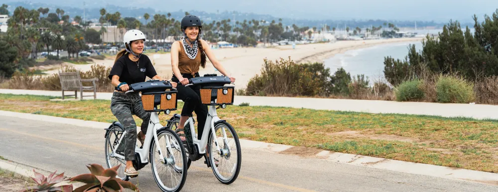 Two women wearing helmets ride BCycle bikes along coast SB